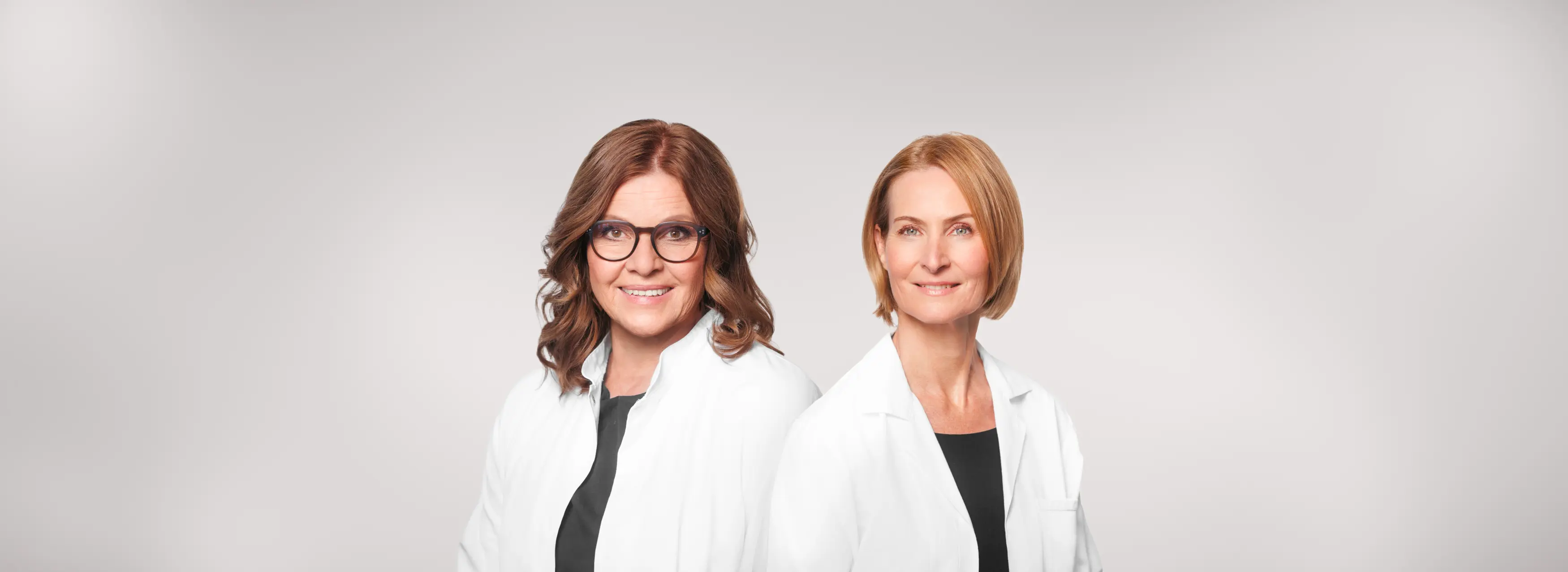 Dr. med. Susanne Spielmann und Dr. med. Christiane Lang-Walker in weißen Kitteln vor hellem Hintergrund, beide lächelnd.
